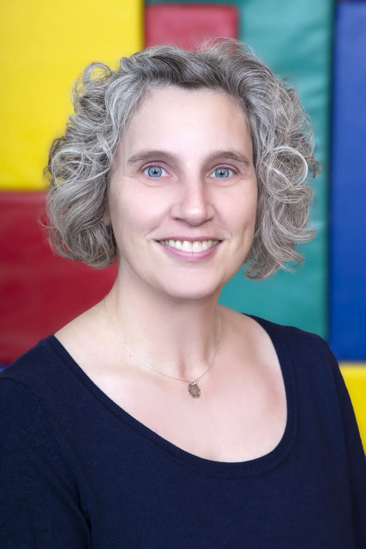 Esther Grunig mit lockigem, grauem Haar und blauer Augen, trägt ein schwarzes Oberteil, vor einem bunten Hintergrund.