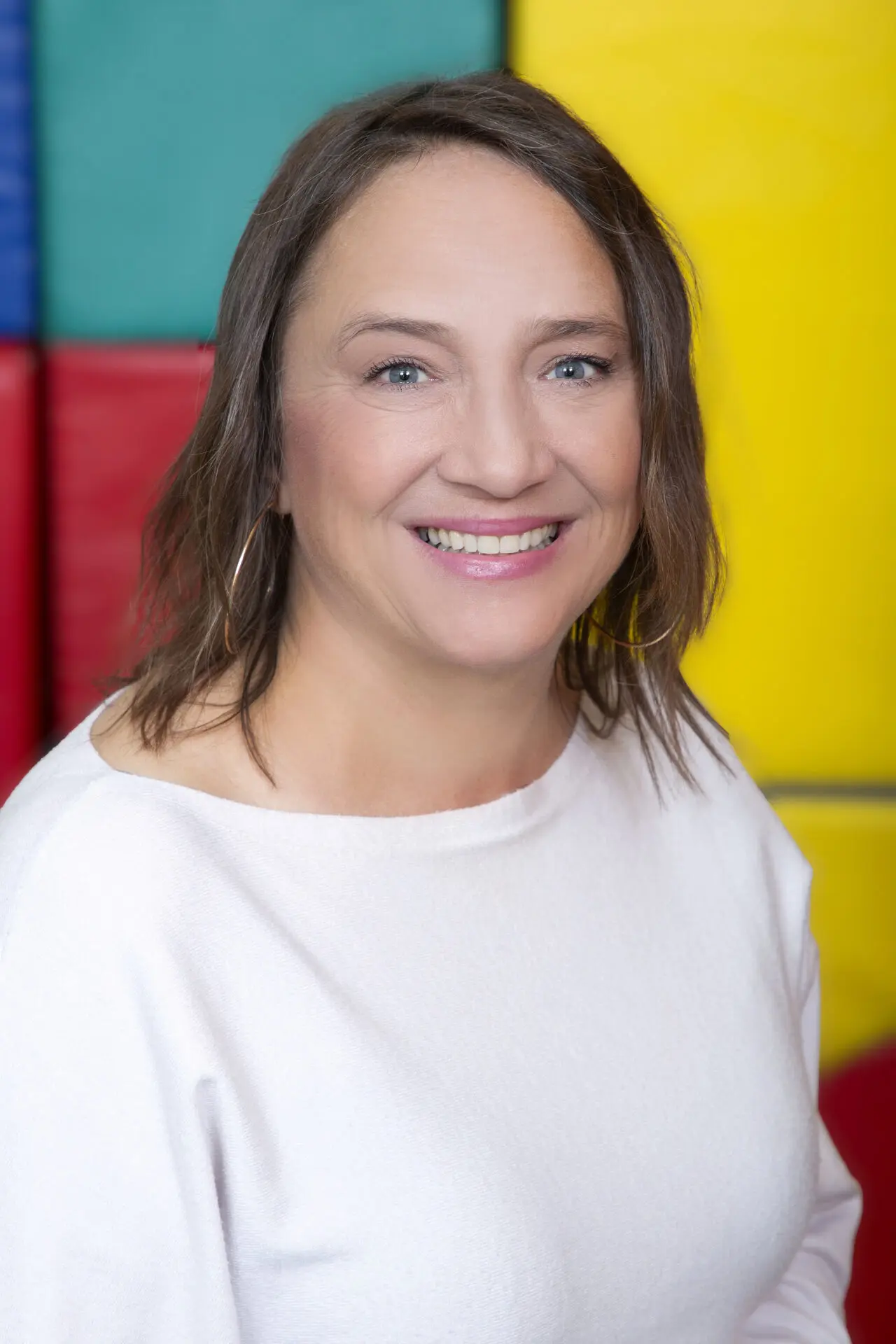 Karin Goj mit lächelndem Gesicht, trägt ein weißes Oberteil, vor bunten Hintergrundelementen in Primärfarben.